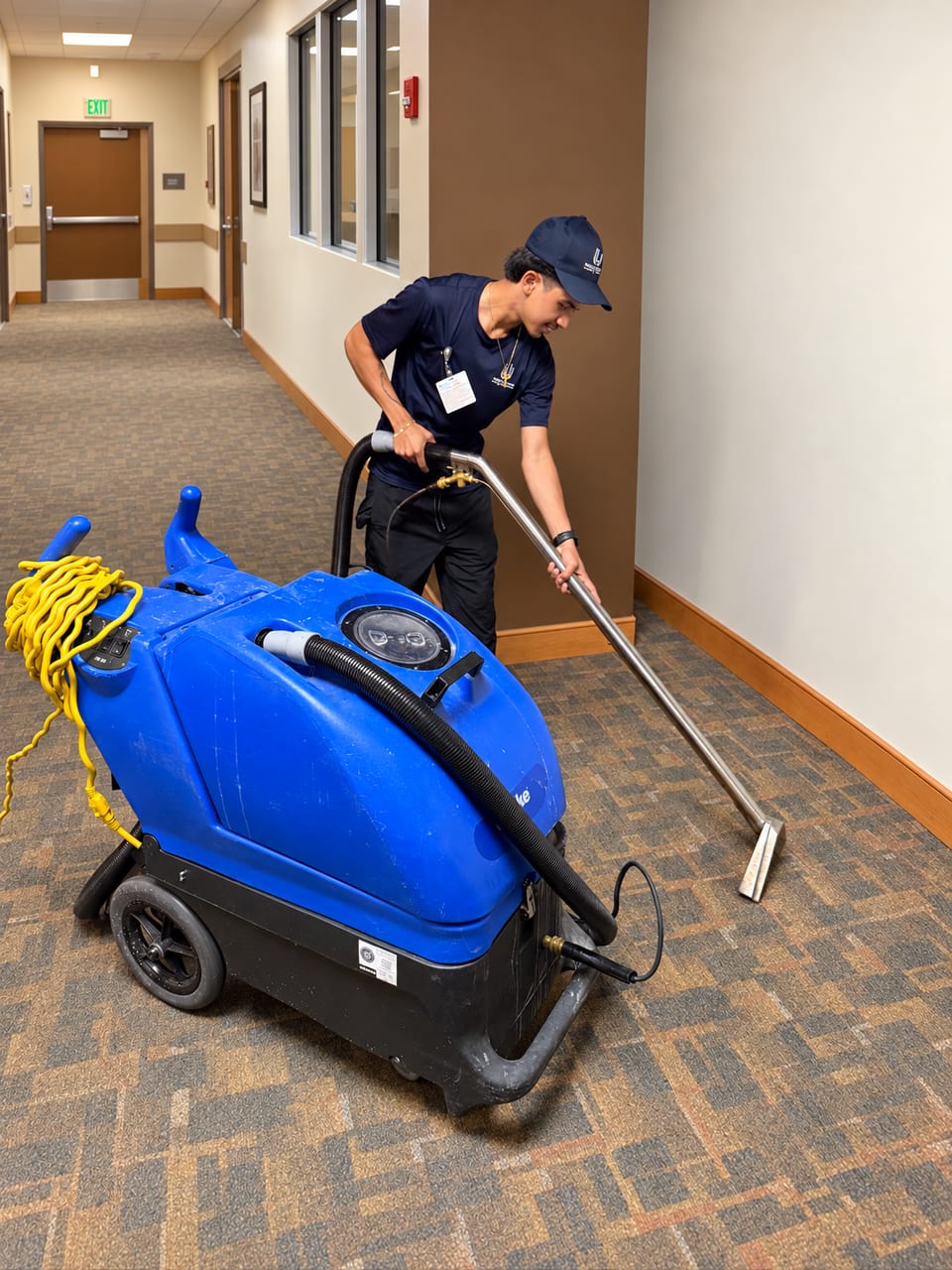 A UFG technician in a navy cap and polo cleans a hospital corridor carpet with an extractor wand and hose from a blue machine