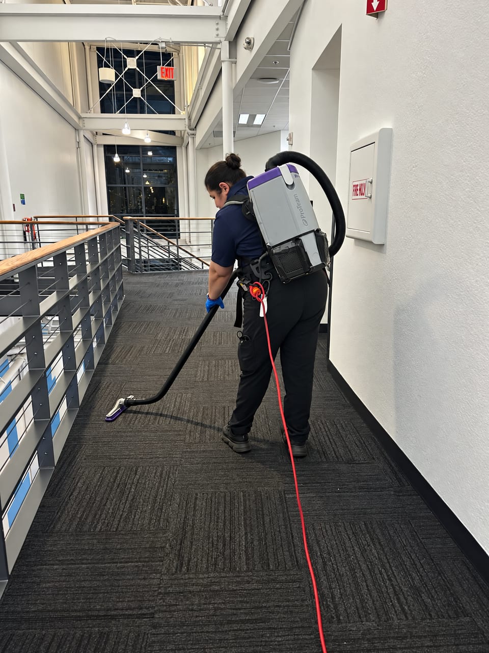 A UFG team member wearing a backpack vacuum walks along a railed museum walkway, cleaning the dark carpet