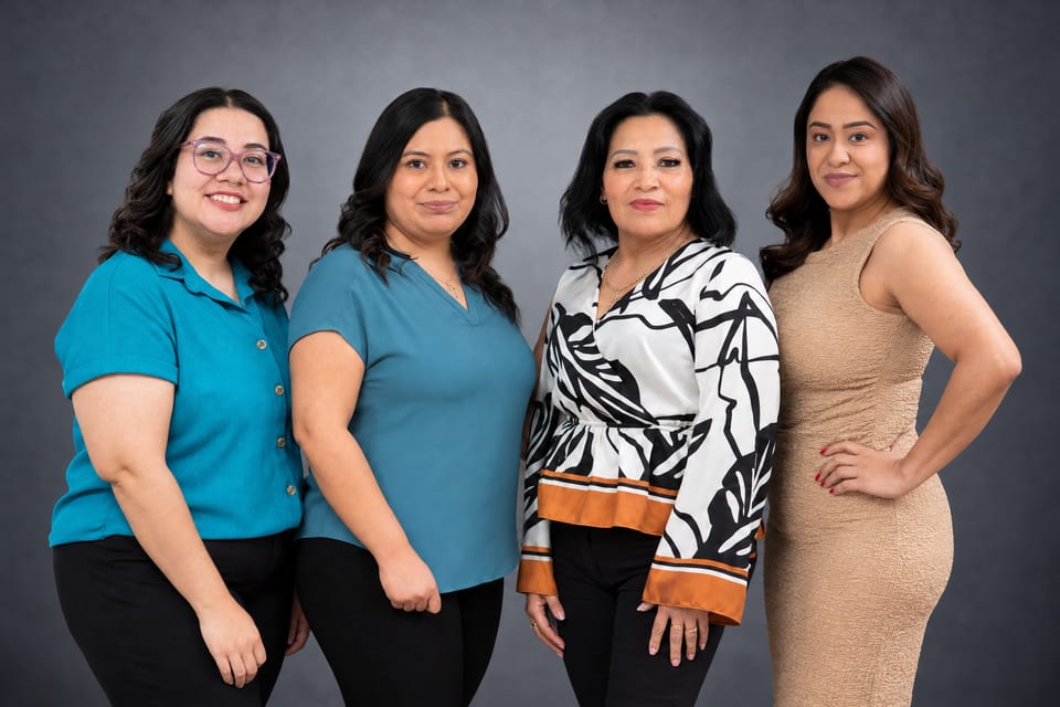 Four women stand side by side in two teal tops, a black-and-white leaf-print blouse and a beige dress on a grey backdrop