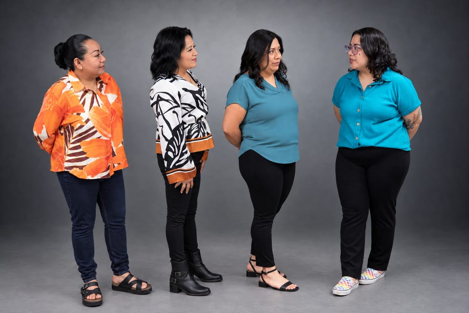 Four women stand in a loose circle facing one another in an orange print shirt, a leaf-print blouse and two teal tops