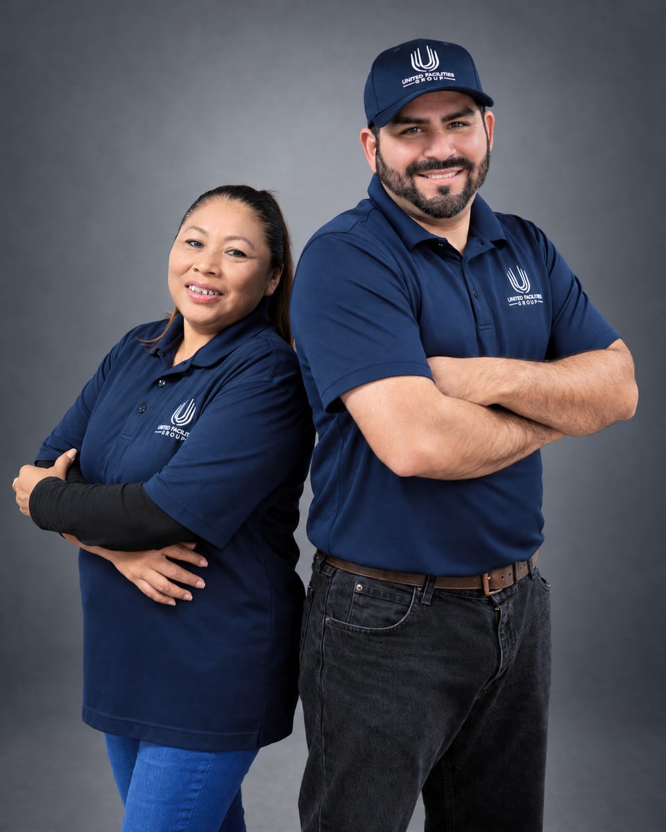 A woman and a bearded man in a UFG cap stand back to back in navy UFG polos, arms folded, smiling on a grey backdrop
