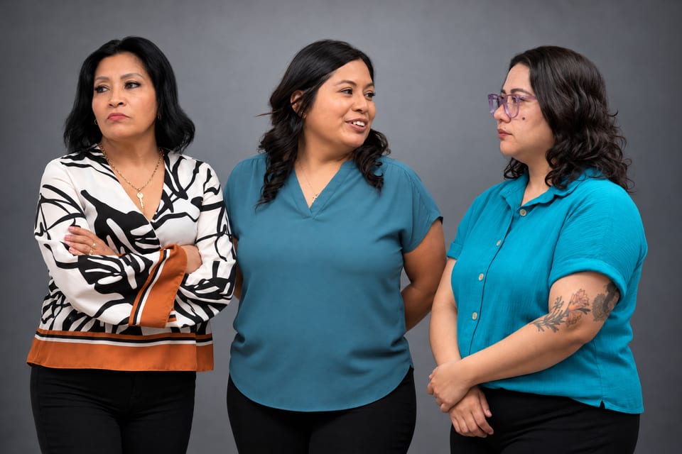 Three women stand close together on a grey backdrop in a leaf-print blouse and two teal tops, looking toward one another