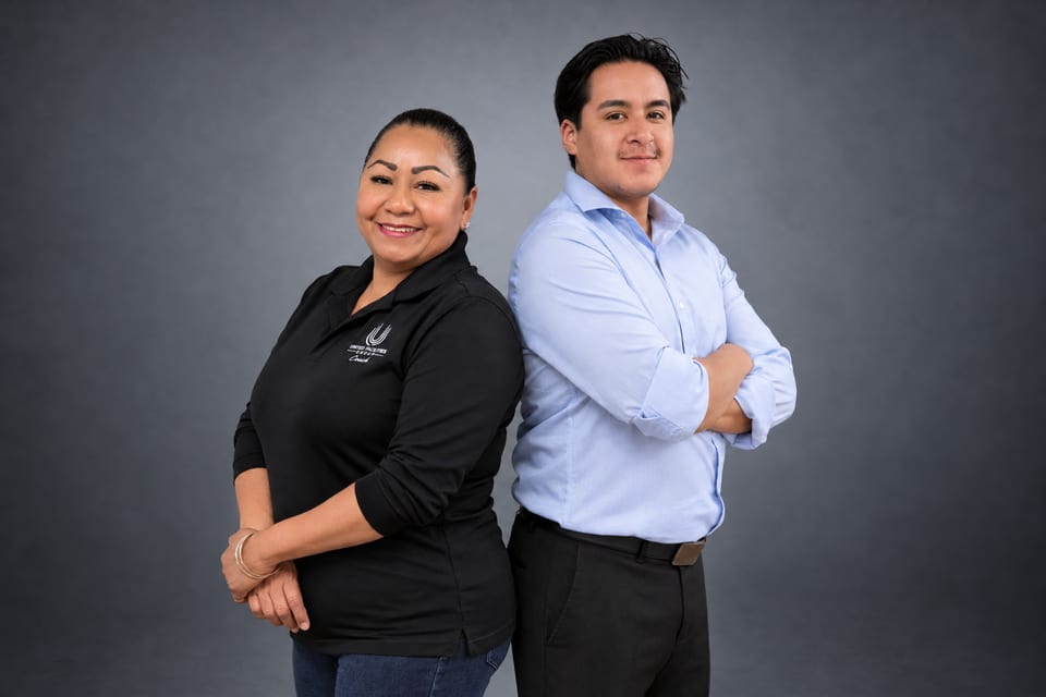 A woman in a black UFG Coach polo and a man in a light-blue shirt stand back to back, he with arms crossed, in a grey studio