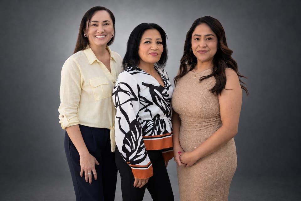 Three women stand close together, smiling, in a pale-yellow shirt, a leaf-print blouse and a beige dress on a grey backdrop