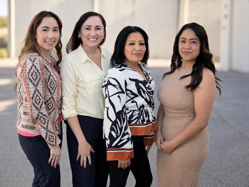 Four women stand close together outdoors before a pale wall, in a patterned cardigan, yellow shirt, print blouse, beige dress