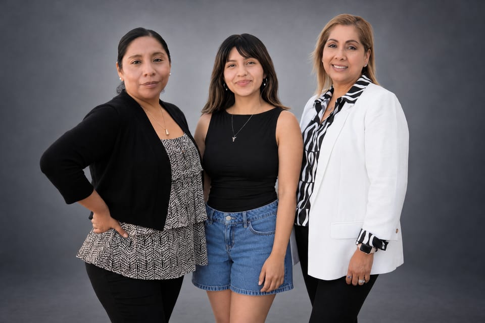 Three women stand together on a grey backdrop in a black cardigan and print top, black with denim shorts, and a white blazer