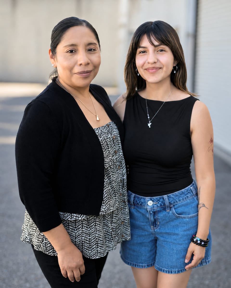 Two women stand close together outdoors, one in a black cardigan over a print top, the other in a black top and denim shorts