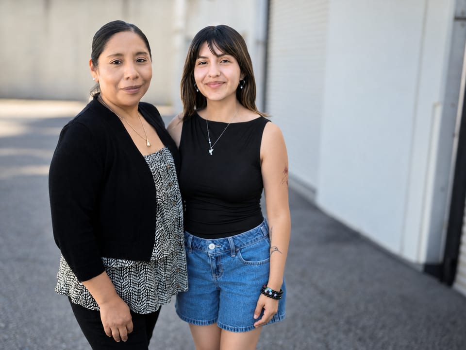 Two women stand close together outdoors, one in a black cardigan over a print top, one in black and denim shorts