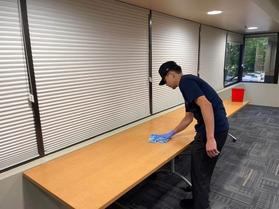 A UFG team member in a black cap and navy shirt wipes a long wooden counter beside a wall of window blinds
