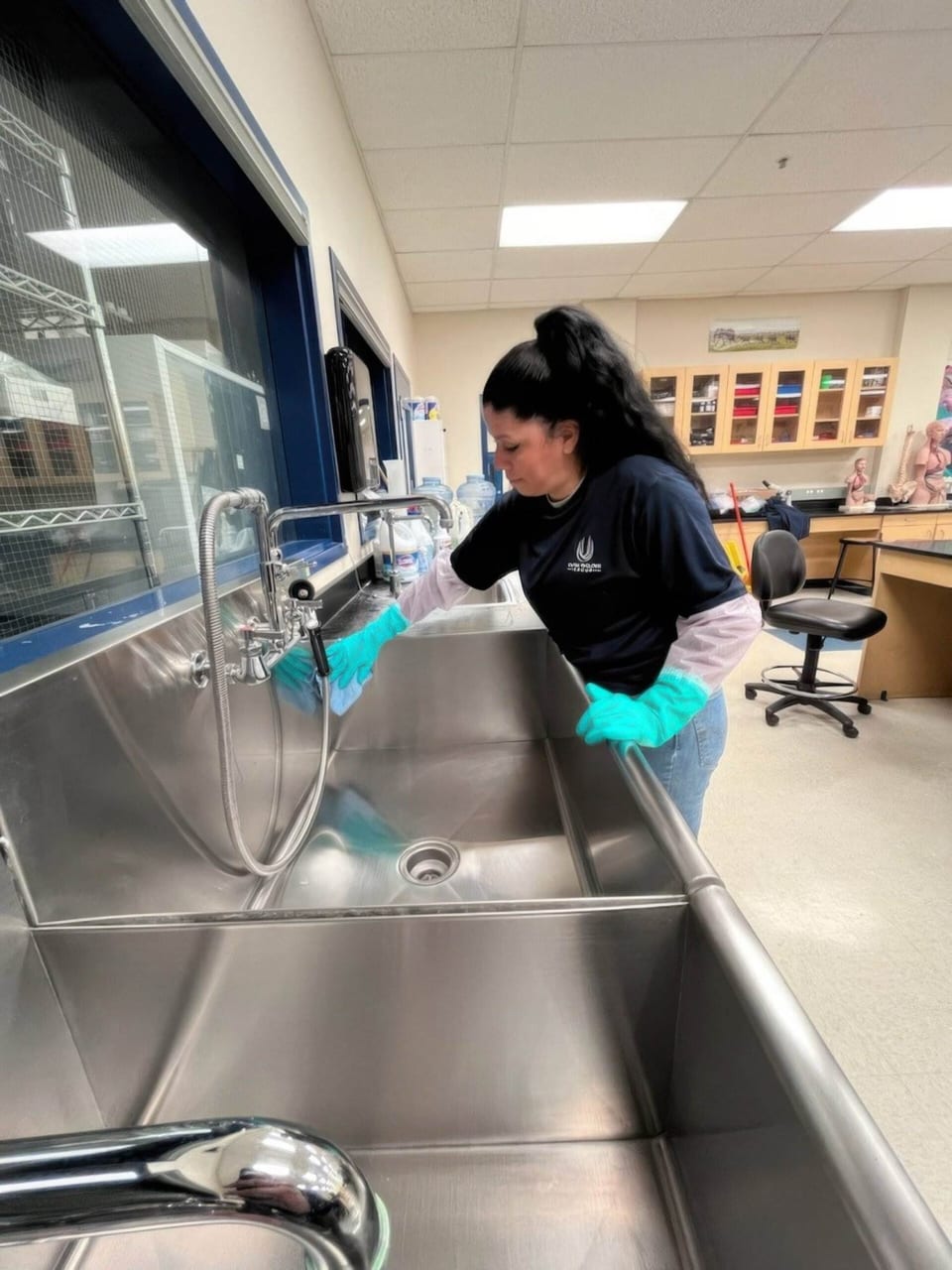 A UFG team member in teal gloves wipes a large stainless-steel sink in a school science lab
