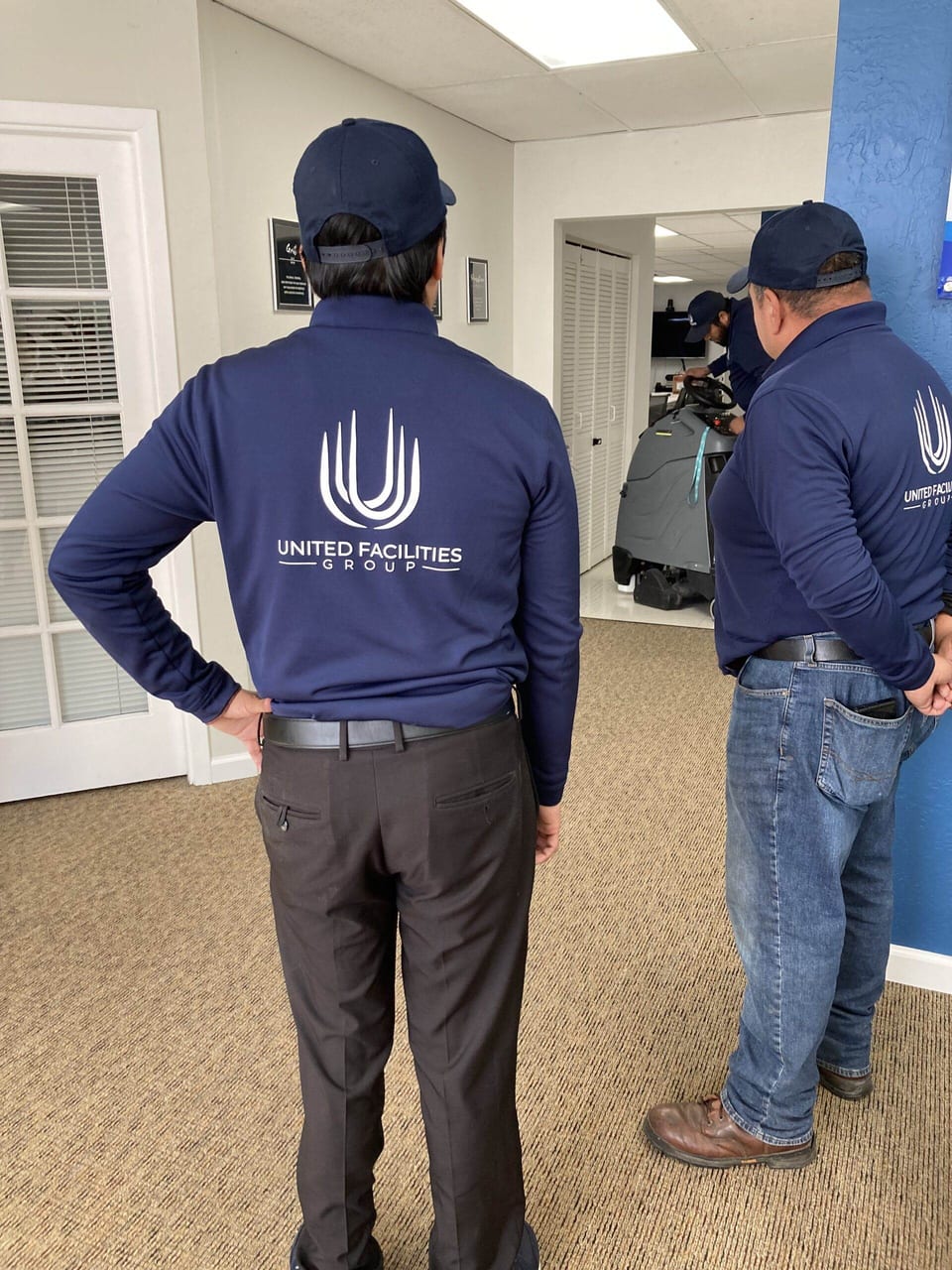 Two UFG team members in navy UFG pullovers and caps, seen from behind, watch a colleague run a floor scrubber down the hall