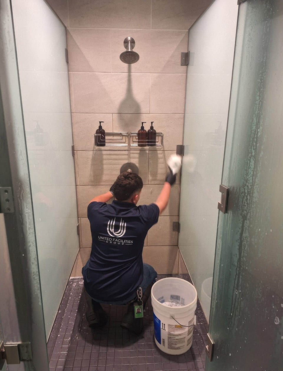 A UFG team member in a navy UFG shirt kneels in a tiled shower stall and scrubs the wall, with a white bucket beside him