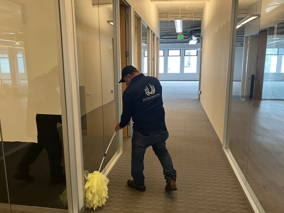 A UFG team member in a navy UFG jacket and cap dust-mops the carpet of a glass-walled office corridor