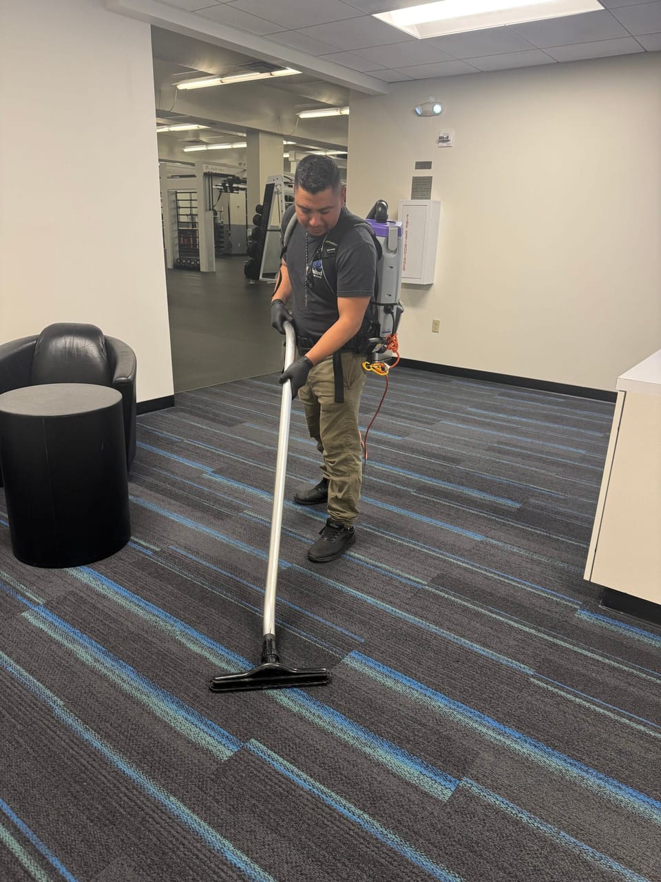 A UFG team member in a grey UFG polo and a backpack vacuum cleans striped carpet in a gym lobby, weight machines behind