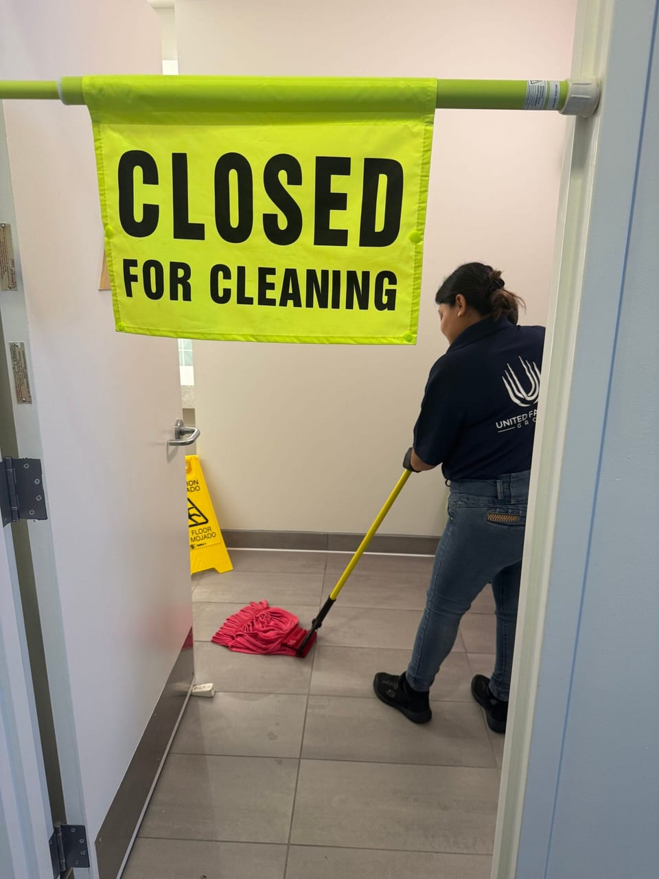 A UFG team member in a navy UFG polo mops a restroom floor with a red mop, a yellow wet-floor cone at the back