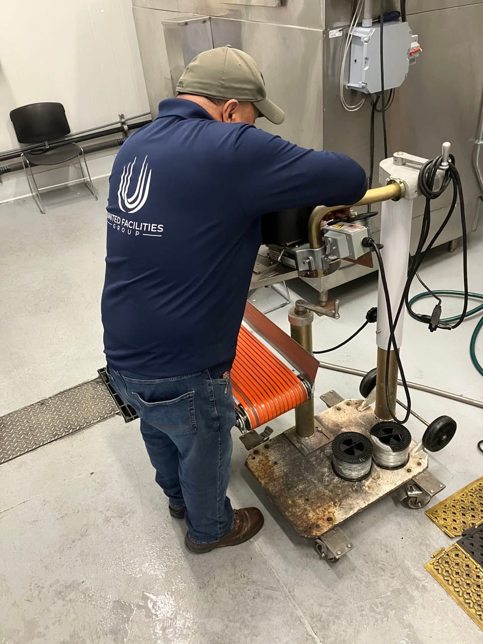 A UFG technician in a navy UFG shirt and khaki cap, seen from behind, works on a machine with an orange belt and tubing