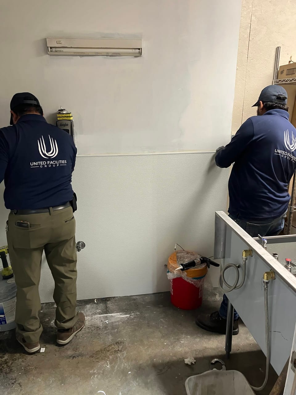 Two UFG technicians in navy UFG shirts, seen from behind, work on a white wall in a bare room beside a sink and red bucket