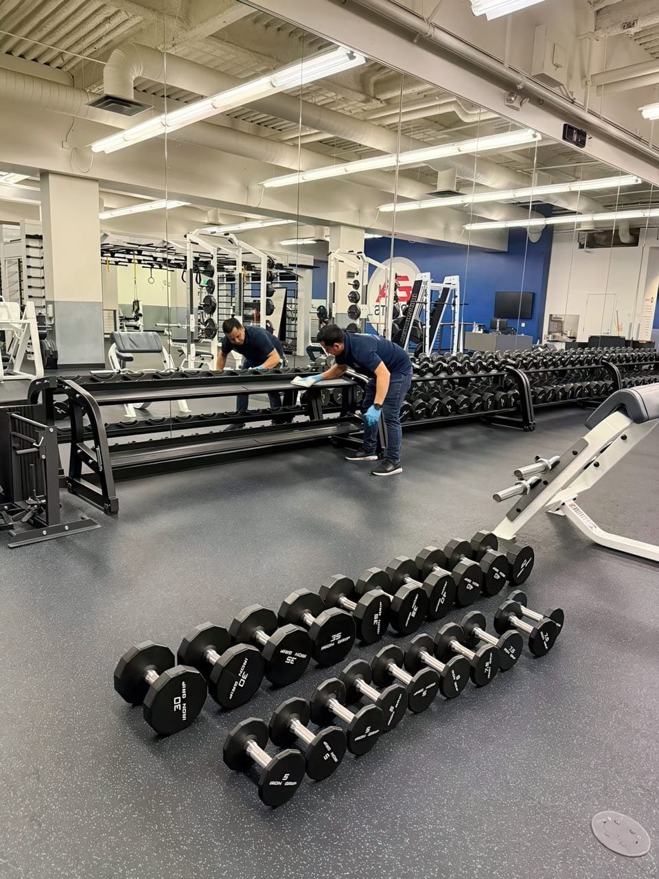 Two UFG team members in navy shirts wipe down dumbbell racks in a company gym with mirrored walls
