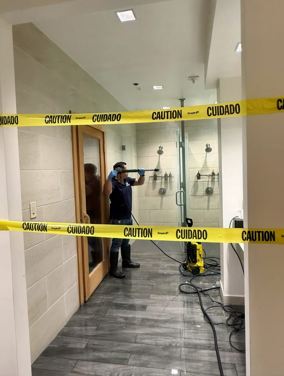 A UFG team member in rubber boots pressure-washes the walls of a tiled shower room, a yellow washer and hose on the floor