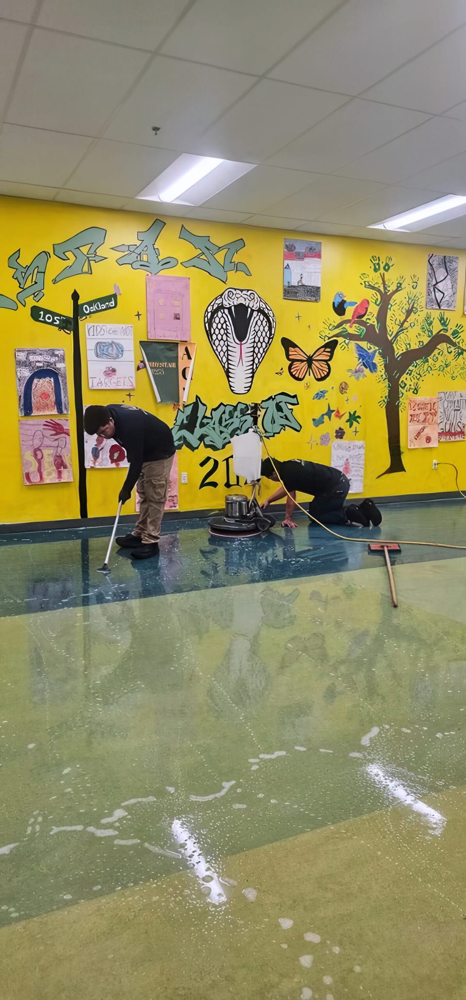Two UFG team members work a rotary floor machine over a wet school floor in front of a yellow student mural