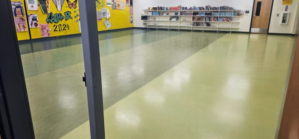 A polished green school floor reflecting the lights, with a bookshelf, a yellow student mural and an open doorway