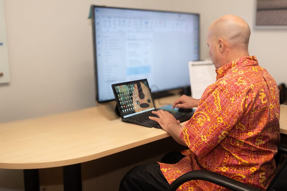 A bald man in a red and gold patterned shirt types on a laptop at a desk in front of a large monitor