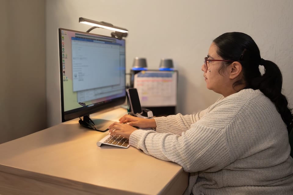 A woman with glasses and a dark ponytail in a cream cardigan types at a computer under a desk lamp