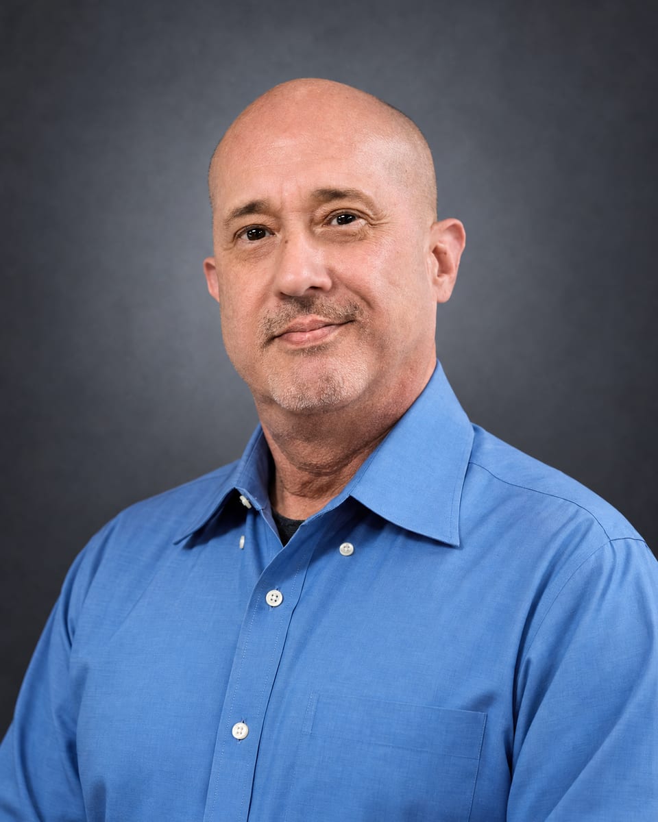 Head-and-shoulders portrait of a bald man with a grey goatee in a blue dress shirt on a grey backdrop